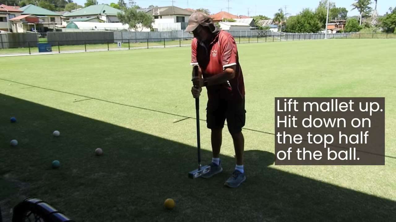 Chris standing over a ball with mallet raised, on-screen caption reading 'Lift mallet up. Hit down on the top half of the ball.'